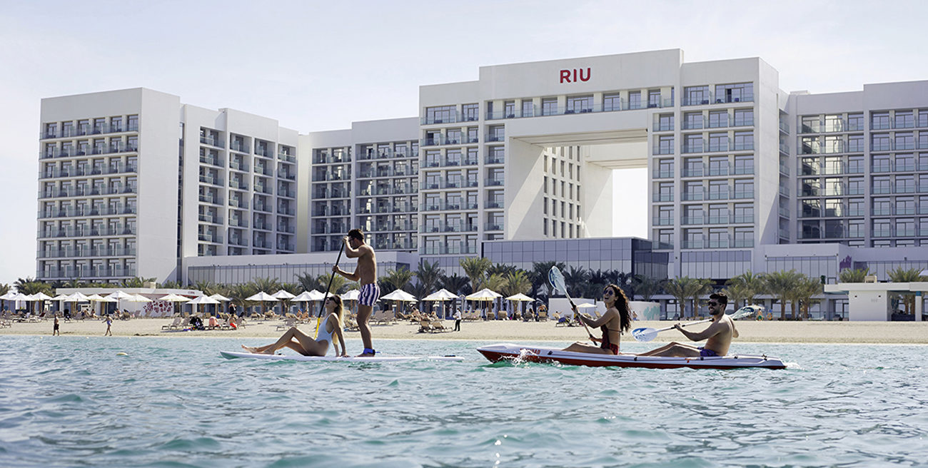 Paddle-boarders in the water, beachfront open-center white resort building in the background labeled RIU