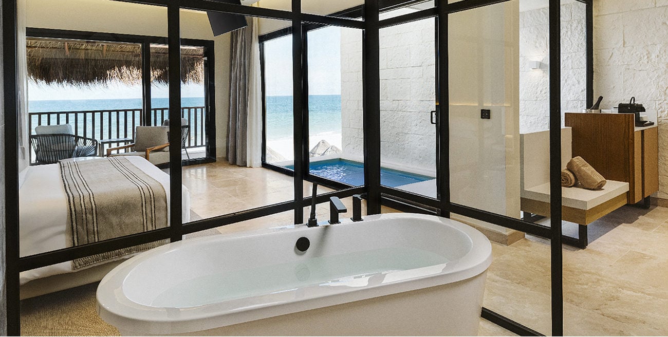 Freestanding tub before glass walls looking out into a private pool, bedroom, and corner of a living area
