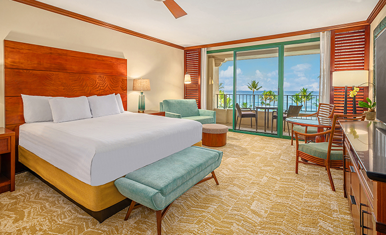 Large bed with warm wood tones and aqua accents in a hotel room with an ocean view balcony