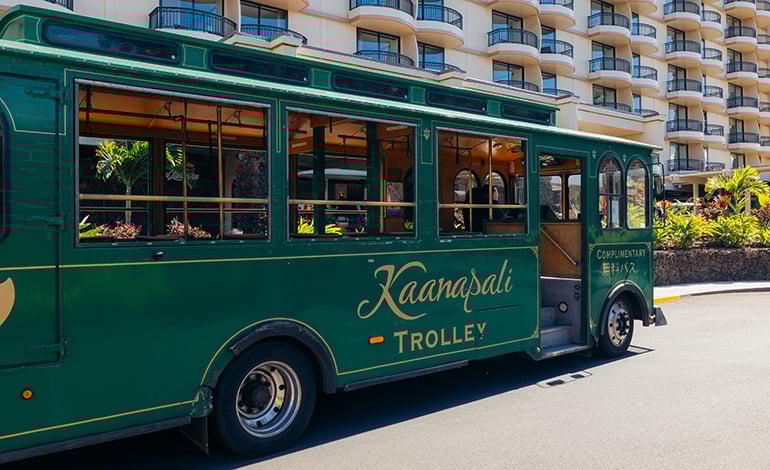 Green trolley on a road before a high-rise residential building labeled Kaanapali Trolley