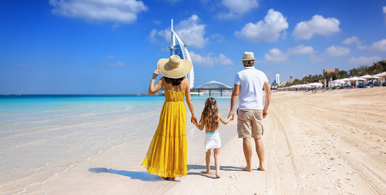 Back view of a woman in a sundress and a man in shorts and a polo shirt on a beach holding hands with a little girl