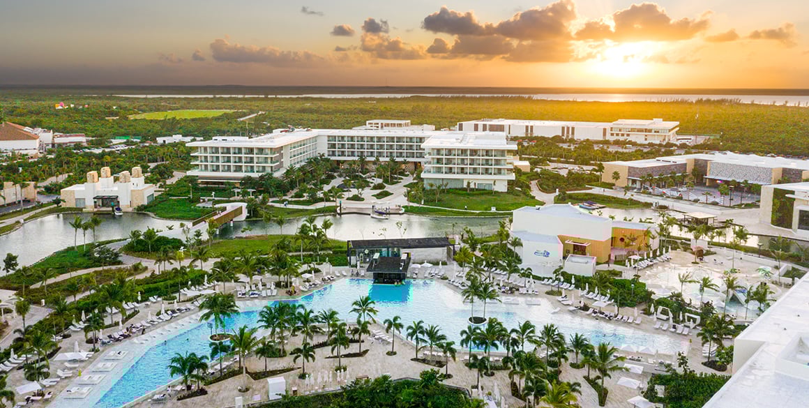 Aerial view of a white resort, giant pool, and mangroves in the background near the setting sun