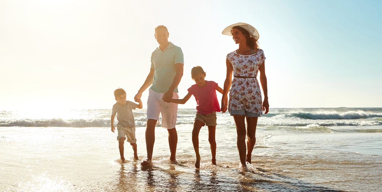 A family with two young children hold hands while walking down the beach.