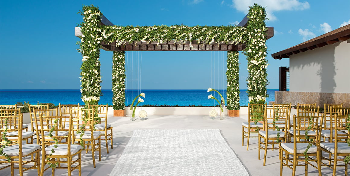 Seaside wedding ceremony setup with a floral pergola, white aisle, and gold chairs overlooking the ocean.