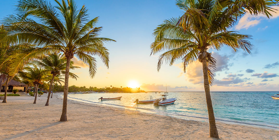 Palm-fringed beach at Akumal Bay during sunset, with small boats floating in the turquoise water.