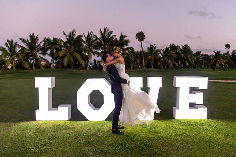 A white wedding couple in front of white letters that spell out love with palm trees in the background
