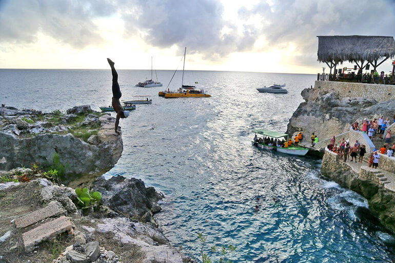 Silhouette of a person dibing off a cliff into water with crowds observing on the opposite shore under a thatched roof