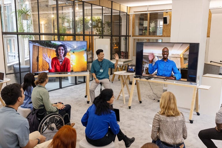 Group of mixed adults sitting on stools in front of a presenter and two people on two TV screens waving