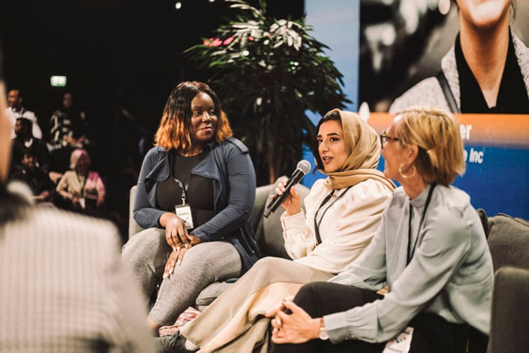 Three multiracial women sitting on stage on a couch as part of a panel before a live audience