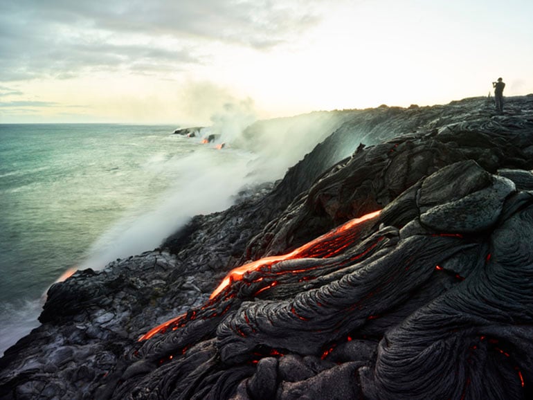 Black rippled lava with red streaks on a cliff with splashing waves and one man's silhouette
