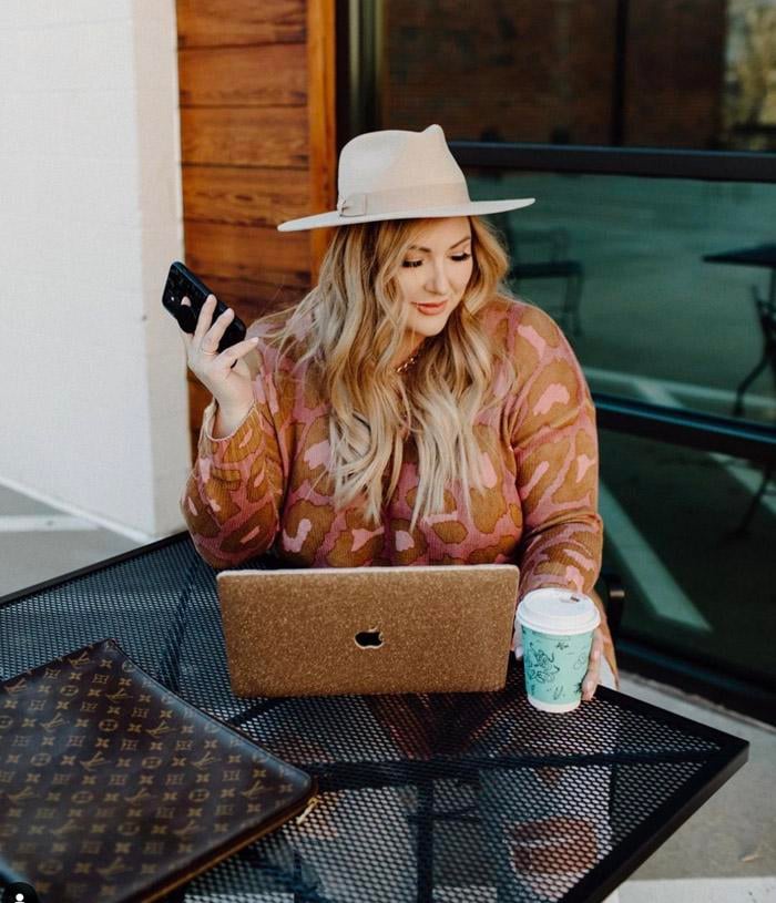 Blonde woman in a camel-colored fedora holding a cell phone and working on a gold-glittered laptop with a cup of coffee to the side
