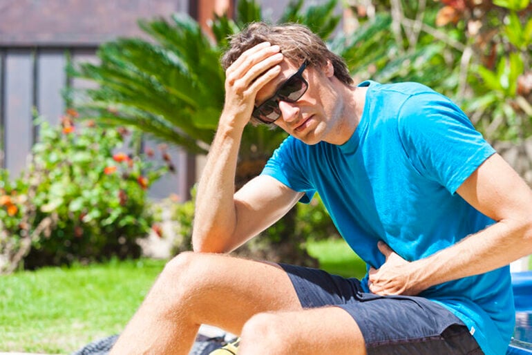 White man in a blue shirt holding stomach in a tropical garden