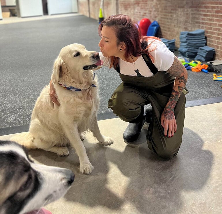 Woman with purple hair kneeling to kiss a golden retriever dog