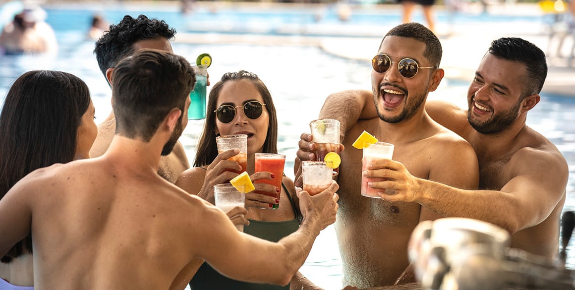 A diverse group of four young men and two women clink cups in a pool 