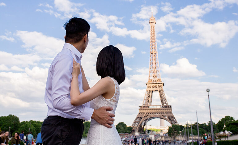 A well-dressed Asian couple embrace while looking at the Eiffel Tower in the distance