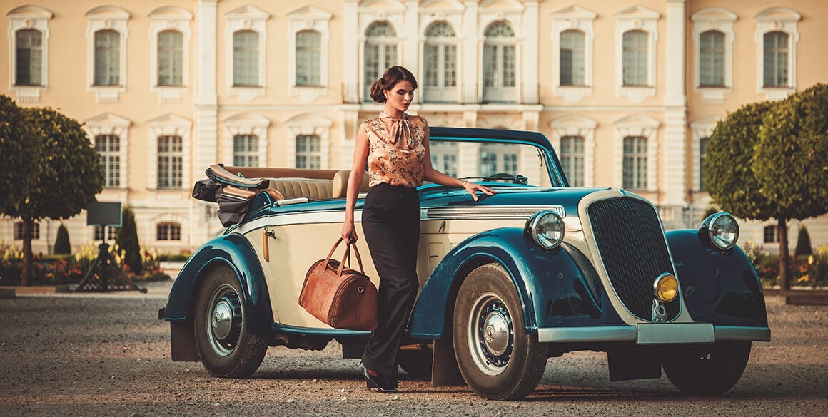 A fashionable young woman stands beside an old-fashioned convertible outside of a historical estate.