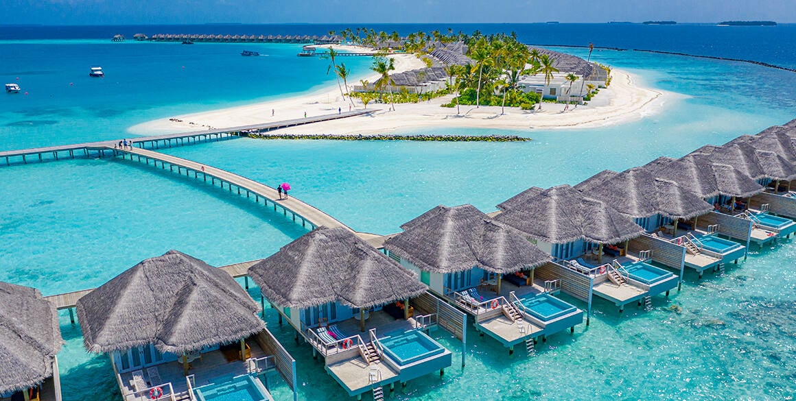 Overwater bungalows in an electric blue sea with a sandy atoll in the background