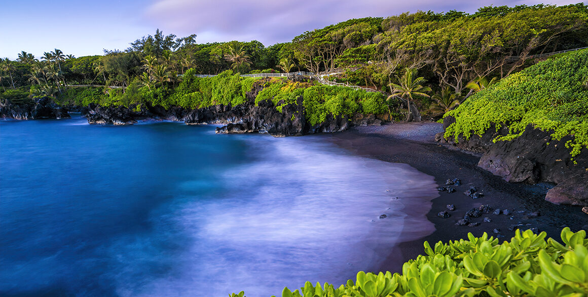 Aerial wide shot of a blue bay with black sand surrounded by greenery