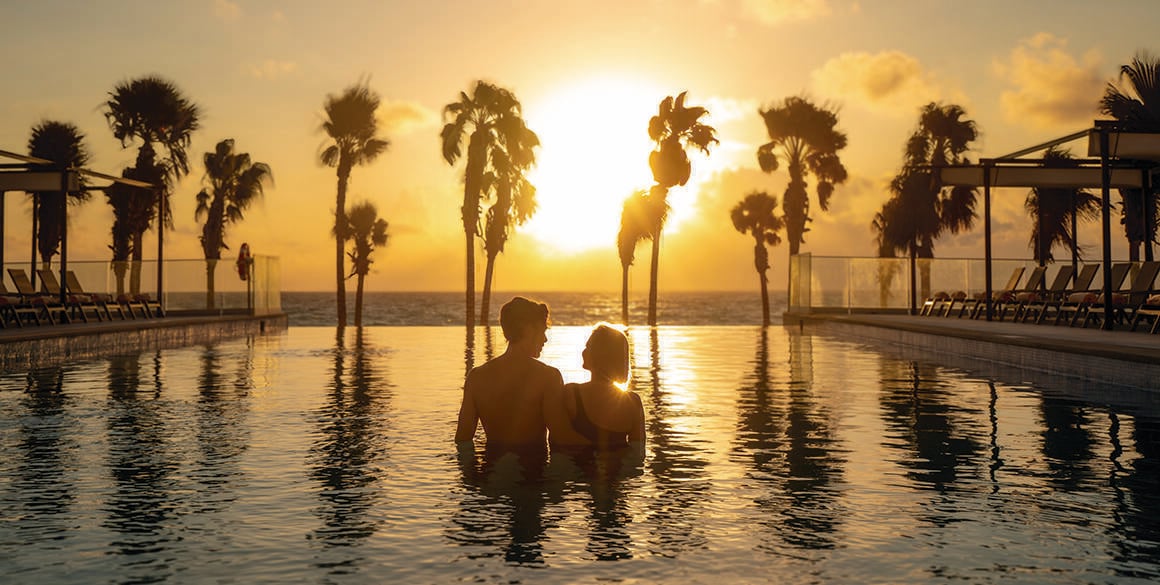 Silhouette of a hetero couple in a pool with the sun lighting up the palm trees