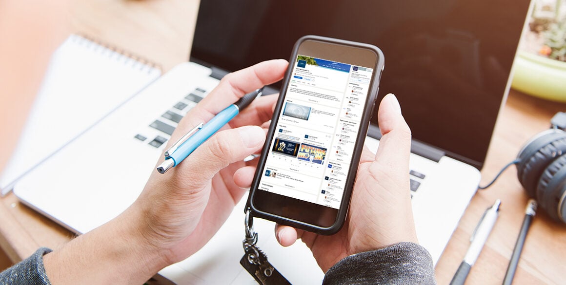 Hands holding a phone with LinkedIn profile up and a blank laptop in the background