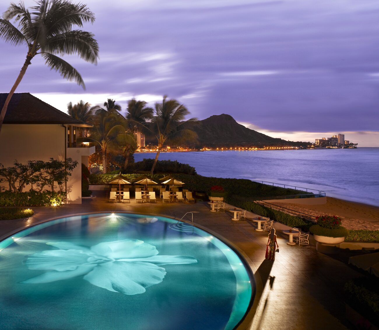 Woman by a pool lit up with a flower in it overlooking the island lit up at night and the ocean