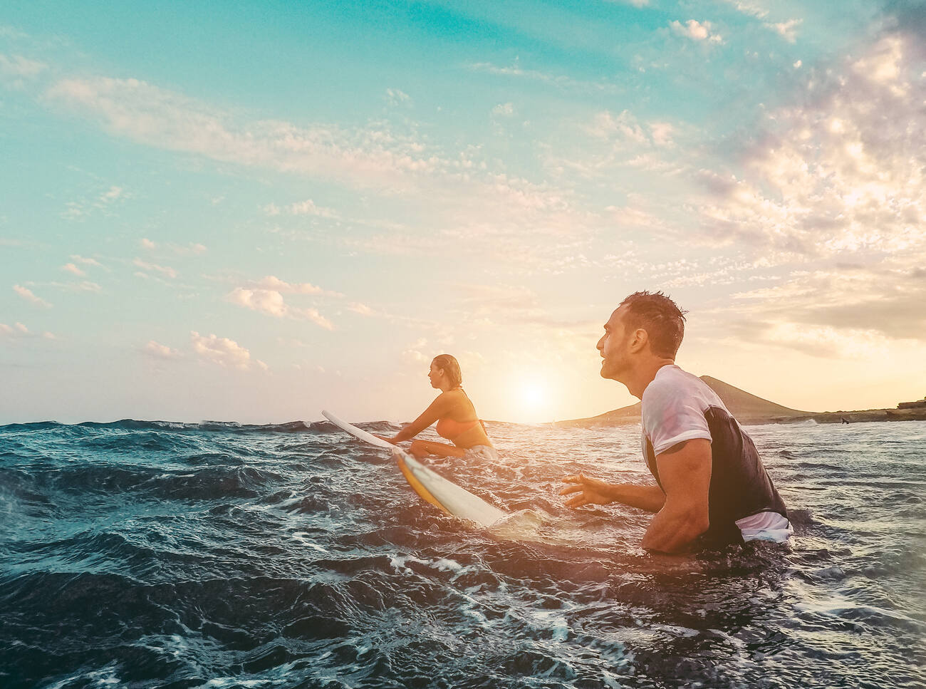 A male and female surfer riding out on their surfboards at sunrise.