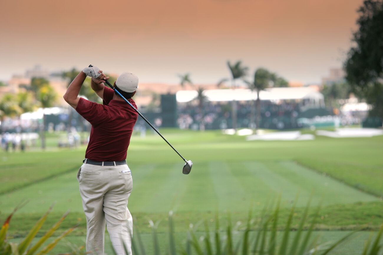 A golfer swings his club on a tropical course at sunset.