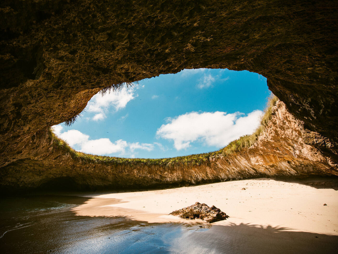 A hidden beach within a cavern. A massive circular opening, like the rim of a crater, looks out toward a blue sky.