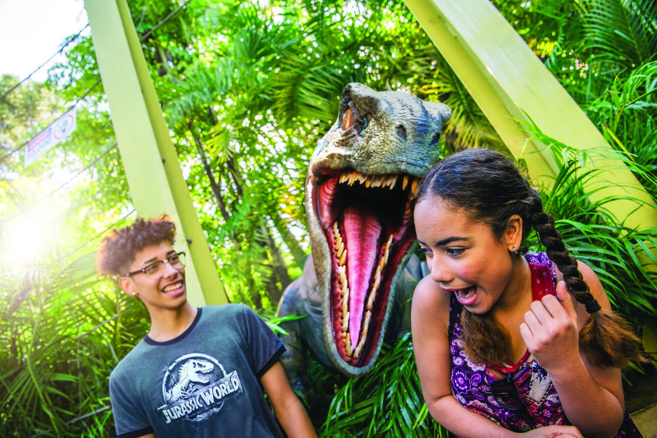 An animatronic velociraptor with its jaws wide open pops up between a laughing boy in a "Jurassic World" t-shirt and a screaming girl at Universal's Islands of Adventure theme park. 