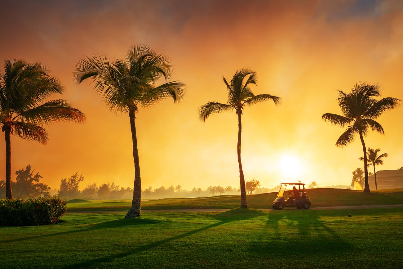 A golf cart drives across a tropical golf course at sunset.