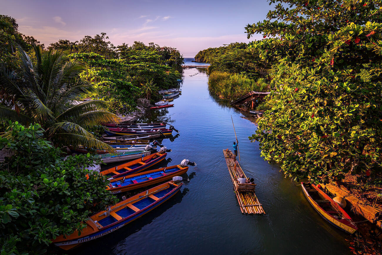 A man steers a bamboo raft with two seated passengers past a row of boats on a river at sunset.