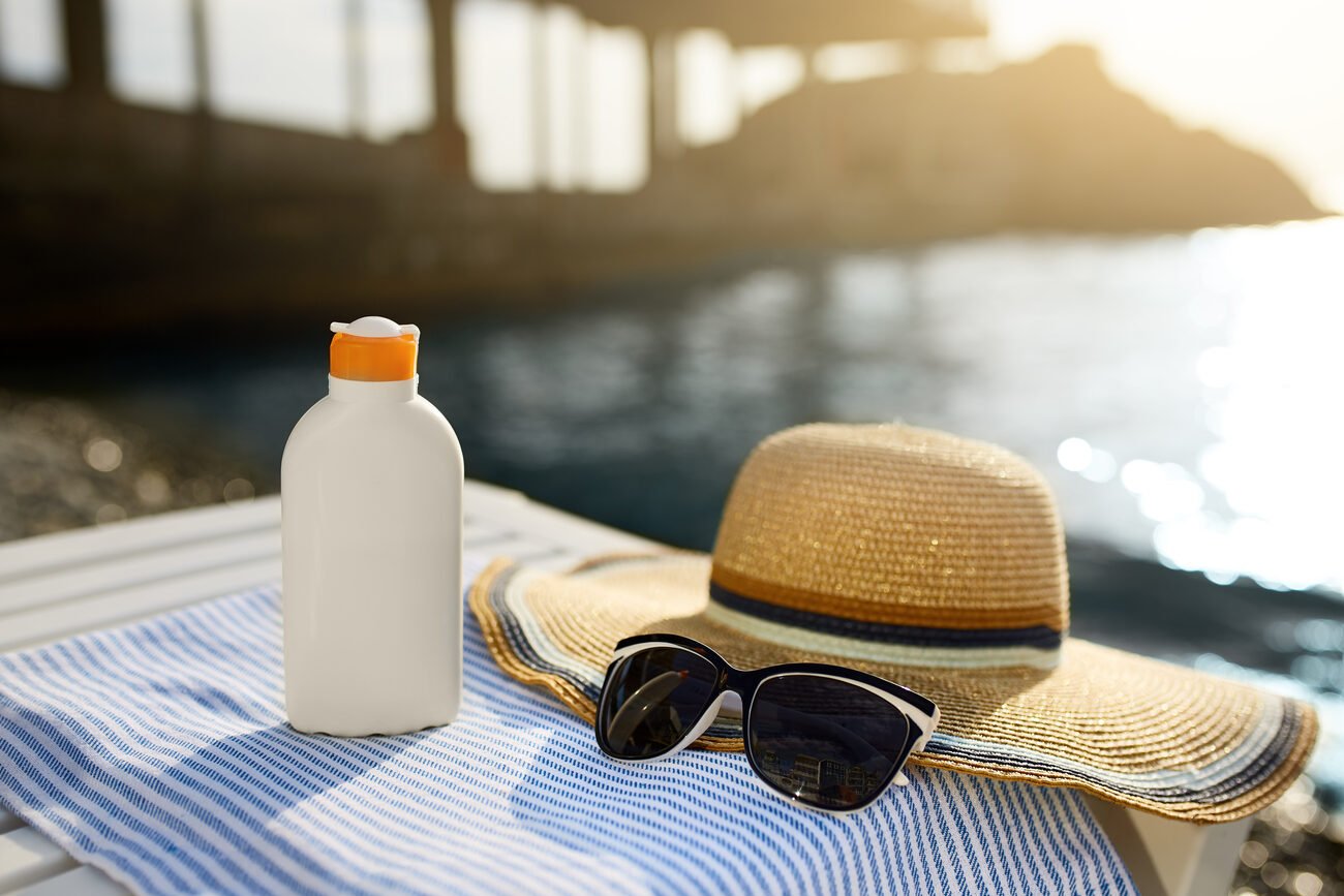 Sunglasses, sunscreen bottle, and sunhat on a blue towel with an ocean pier in the background.