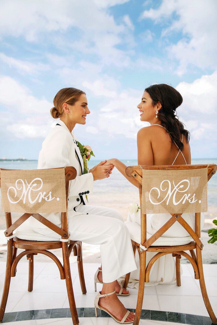 Brides sitting in 'Mrs.' chairs on the beach