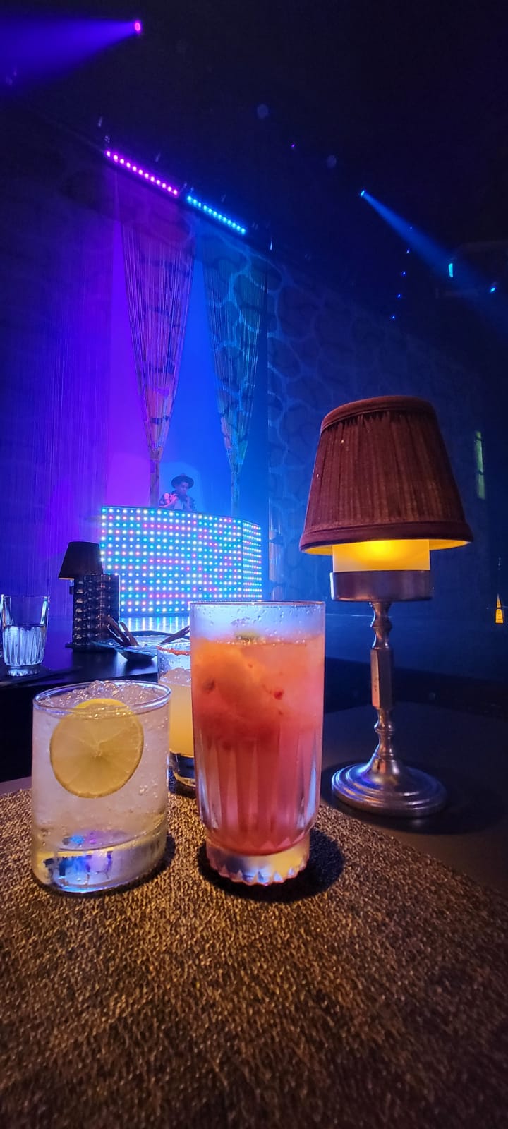 Two drinks on a table with a DJ playing in the back
