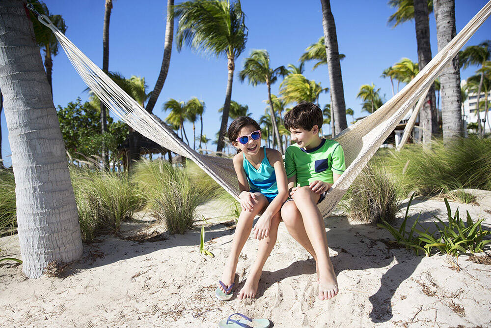 A boy and a girl sitting on a hammock strung between two palm trees on Palm Beach.